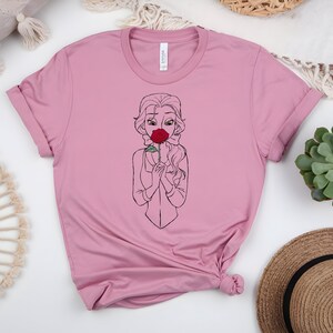 May include: A light pink t-shirt featuring a line drawing of a woman holding a red rose to her face. The woman has long wavy hair and is wearing a blazer. The shirt is made of a soft material and has a crew neck.