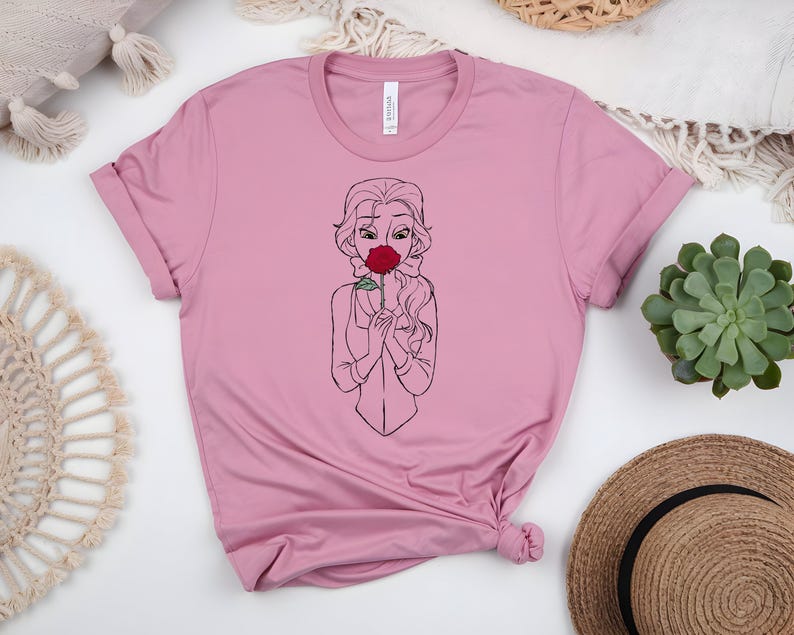 May include: A light pink t-shirt featuring a line drawing of a woman holding a red rose to her face. The woman has long wavy hair and is wearing a blazer. The shirt is made of a soft material and has a crew neck.