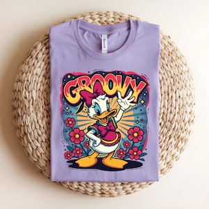 May include: A lilac-colored t-shirt featuring a cartoon graphic of a duck with a pink bow and the word "GROOVY" in a colorful, retro font. The design includes flowers and radiating lines, creating a vibrant, vintage aesthetic. The shirt is laid flat.