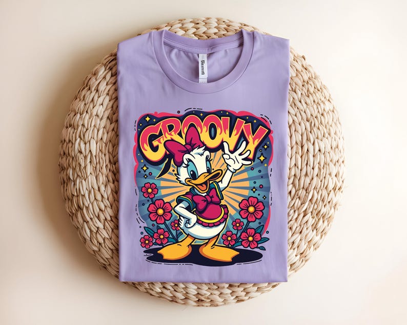 May include: A lilac-colored t-shirt featuring a cartoon graphic of a duck with a pink bow and the word "GROOVY" in a colorful, retro font. The design includes flowers and radiating lines, creating a vibrant, vintage aesthetic. The shirt is laid flat.