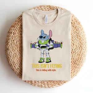 May include: Beige t-shirt featuring a cartoon character, Stitch, dressed as Buzz Lightyear with the text "THIS ISN'T FLYING" in yellow. The shirt has a crew neck and is folded on a woven mat.
