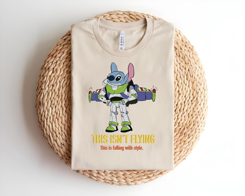 May include: Beige t-shirt featuring a cartoon character, Stitch, dressed as Buzz Lightyear with the text "THIS ISN'T FLYING" in yellow. The shirt has a crew neck and is folded on a woven mat.