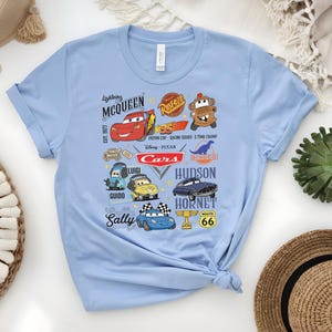 May include: Light blue t-shirt featuring a colorful graphic of characters and logos from the Disney Pixar Cars movie. The design includes Lightning McQueen, Mater, and other characters, along with the text "Cars" and "Route 66".