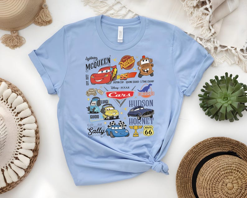 May include: Light blue t-shirt featuring a colorful graphic of characters and logos from the Disney Pixar Cars movie. The design includes Lightning McQueen, Mater, and other characters, along with the text "Cars" and "Route 66".
