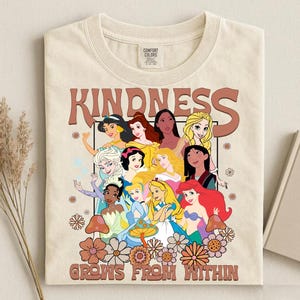 May include: Cream-colored t-shirt featuring a graphic of Disney princesses with the word "KINDNESS" above and "GROWS FROM WITHIN" below. The design includes flowers and mushrooms in shades of brown and orange.