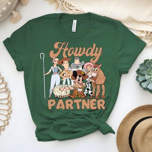May include: Green t-shirt featuring a cartoon graphic of characters with the text "Howdy Partner". The graphic includes a shepherdess, Woody, Buzz Lightyear, Jessie, a horse, and Mickey Mouse in a cowboy outfit. The shirt is a crew neck.