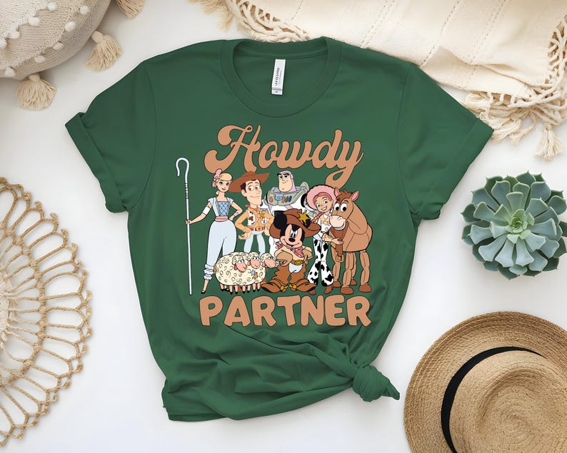May include: Green t-shirt featuring a cartoon graphic of characters with the text "Howdy Partner". The graphic includes a shepherdess, Woody, Buzz Lightyear, Jessie, a horse, and Mickey Mouse in a cowboy outfit. The shirt is a crew neck.