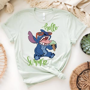K&ouml;nnte beinhalten: Hellgr&uuml;nes T-Shirt mit einer Comicfigur, die eine Sonnenbrille tr&auml;gt und eine Eist&uuml;te h&auml;lt. Die W&ouml;rter "hello" und "Summer" sind in gr&uuml;ner Schrift geschrieben. Die Figur ist blau mit einem gro&szlig;en Mund und einer rosa Zunge.