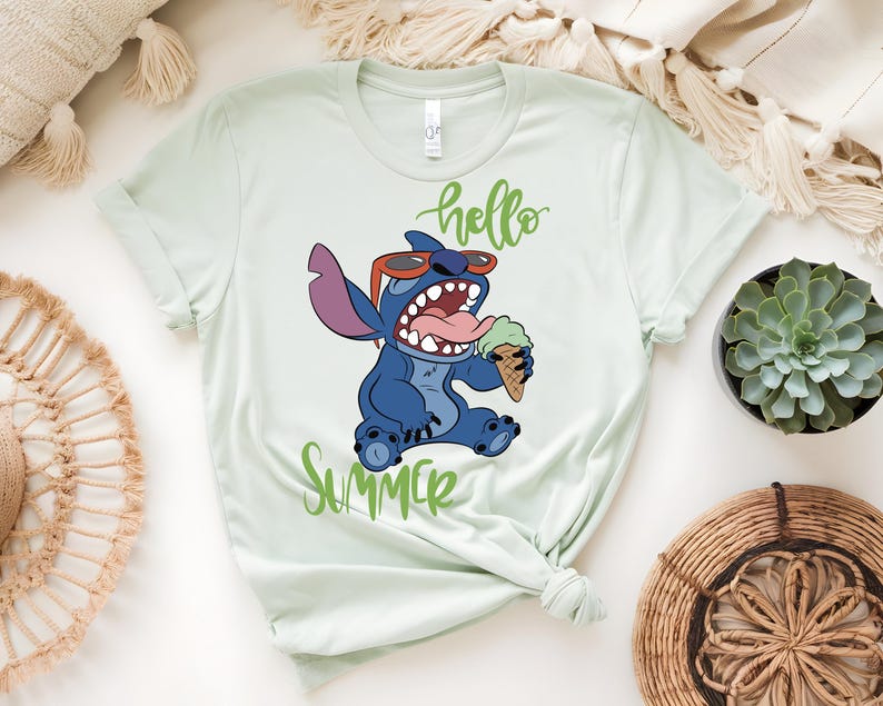 K&ouml;nnte beinhalten: Hellgr&uuml;nes T-Shirt mit einer Comicfigur, die eine Sonnenbrille tr&auml;gt und eine Eist&uuml;te h&auml;lt. Die W&ouml;rter "hello" und "Summer" sind in gr&uuml;ner Schrift geschrieben. Die Figur ist blau mit einem gro&szlig;en Mund und einer rosa Zunge.