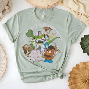 May include: Pale green t-shirt with a graphic of Toy Story characters. The design includes Woody, Buzz Lightyear, Rex, Slinky Dog, Hamm, and Mr. Potato Head, with the text "YOU'VE GOT A FRIEND IN ME" in a circular design.