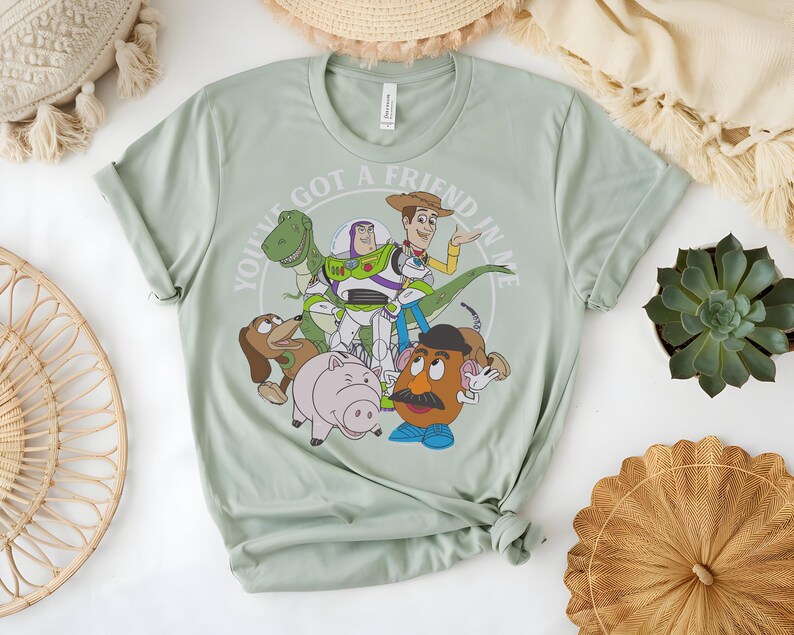 May include: Pale green t-shirt with a graphic of Toy Story characters. The design includes Woody, Buzz Lightyear, Rex, Slinky Dog, Hamm, and Mr. Potato Head, with the text "YOU'VE GOT A FRIEND IN ME" in a circular design.
