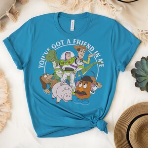 May include: A turquoise t-shirt with a graphic of Toy Story characters. The design includes Woody, Buzz Lightyear, Rex, Mr. Potato Head, Hamm, and Slinky Dog, with the text "YOU'VE GOT A FRIEND IN ME" in a white circle.