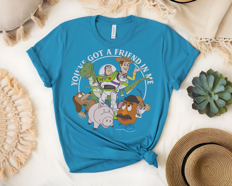 May include: A turquoise t-shirt with a graphic of Toy Story characters. The design includes Woody, Buzz Lightyear, Rex, Mr. Potato Head, Hamm, and Slinky Dog, with the text "YOU'VE GOT A FRIEND IN ME" in a white circle.