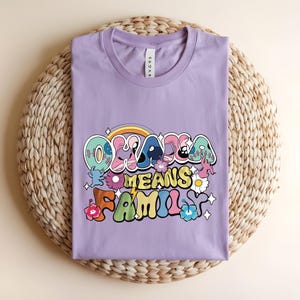 Puede incluir: Una camiseta lila con la frase "Oklahoma Means Family" en una fuente retro colorida. El dise&ntilde;o incluye un arco&iacute;ris, flores y estrellas. La camiseta est&aacute; doblada sobre una estera redonda tejida.