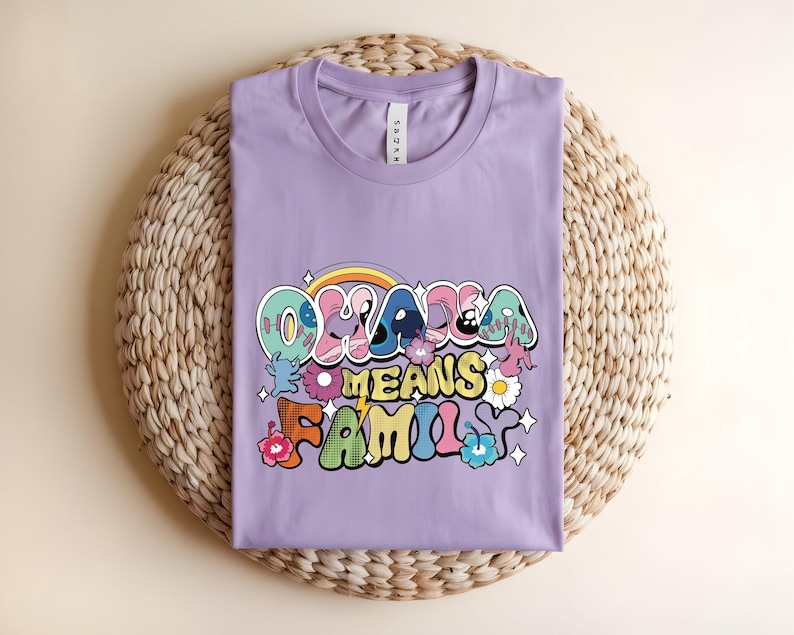 Puede incluir: Una camiseta lila con la frase "Oklahoma Means Family" en una fuente retro colorida. El dise&ntilde;o incluye un arco&iacute;ris, flores y estrellas. La camiseta est&aacute; doblada sobre una estera redonda tejida.