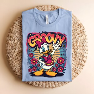 May include: A light blue t-shirt featuring a cartoon of a white duck with a pink bow and dress, surrounded by flowers. The word "GROOVY" is written above the duck in a colorful, retro font.