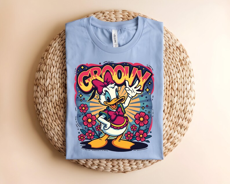 May include: A light blue t-shirt featuring a cartoon of a white duck with a pink bow and dress, surrounded by flowers. The word "GROOVY" is written above the duck in a colorful, retro font.