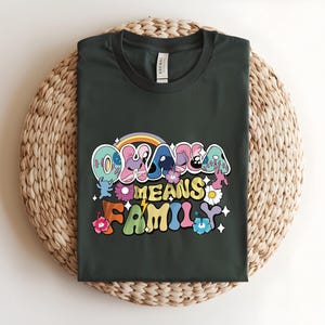 Puede incluir: Camiseta verde oscuro con la frase "OHANA MEANS FAMILY" en una fuente retro colorida. El dise&ntilde;o incluye un arco&iacute;ris, flores y estrellas. La camiseta se muestra sobre una estera redonda tejida.