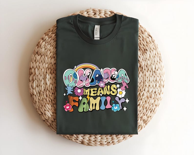 Puede incluir: Camiseta verde oscuro con la frase "OHANA MEANS FAMILY" en una fuente retro colorida. El dise&ntilde;o incluye un arco&iacute;ris, flores y estrellas. La camiseta se muestra sobre una estera redonda tejida.