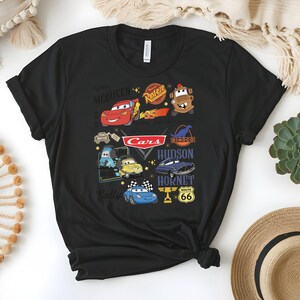 May include: Black t-shirt featuring a colorful graphic of characters and logos from the animated movie "Cars." The design includes Lightning McQueen, Mater, and other characters, along with the "Cars" logo and Route 66 signage. The shirt is made of a soft material.