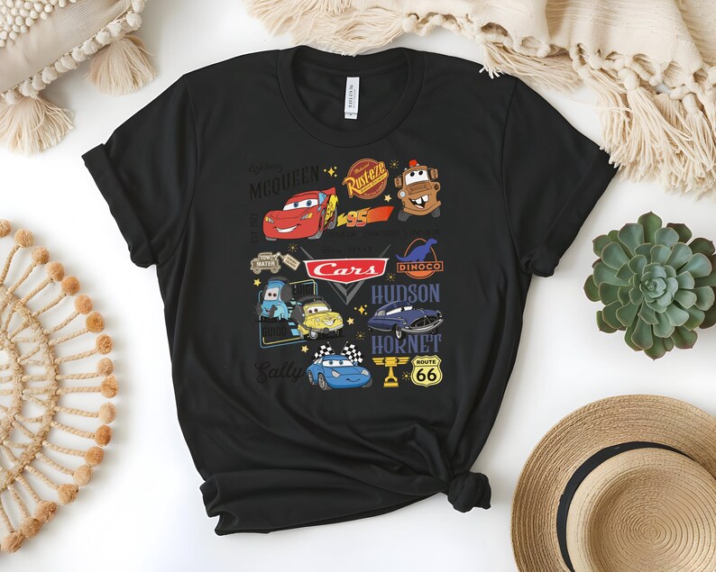 May include: Black t-shirt featuring a colorful graphic of characters and logos from the animated movie "Cars." The design includes Lightning McQueen, Mater, and other characters, along with the "Cars" logo and Route 66 signage. The shirt is made of a soft material.