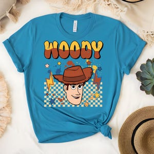 Puede incluir: Una camiseta turquesa con un dibujo animado de Woody de Toy Story. La palabra "WOODY" est&aacute; en letras marrones y amarillas sobre la cara de Woody, con estrellas, rayos y un patr&oacute;n de cuadros.