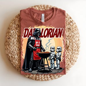 May include: A terracotta t-shirt with a graphic of Darth Vader wearing an apron that says "#1 Dad" grilling with two Stormtroopers. The text "Darthlorian" is above the image.