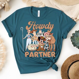 May include: Teal t-shirt featuring a cartoon graphic of characters with the text "Howdy Partner" in a retro font. The graphic includes a shepherdess, Woody, Buzz Lightyear, Jessie, a horse, and Mickey Mouse.