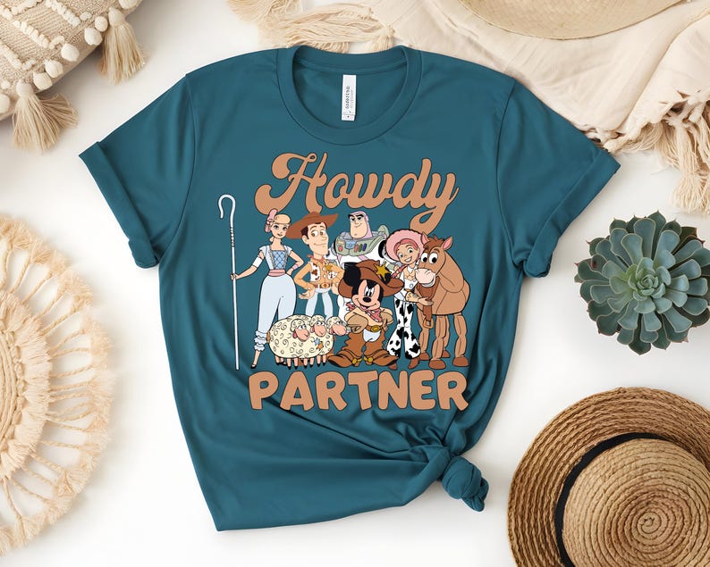 May include: Teal t-shirt featuring a cartoon graphic of characters with the text "Howdy Partner" in a retro font. The graphic includes a shepherdess, Woody, Buzz Lightyear, Jessie, a horse, and Mickey Mouse.