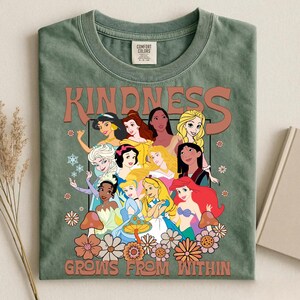 May include: Green Comfort Colors t-shirt featuring a graphic of various cartoon princesses. The word "KINDNESS" is printed above the princesses, and "GROWS FROM WITHIN" is printed below. The design includes flowers and snowflakes.