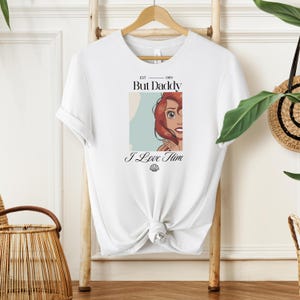 May include: White t-shirt with a graphic of a red-haired cartoon character and the text "But Daddy I Love Him" in black. The shirt is knotted at the waist and hangs on a wooden hanger.