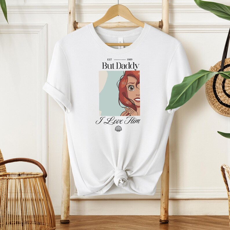 May include: White t-shirt with a graphic of a red-haired cartoon character and the text "But Daddy I Love Him" in black. The shirt is knotted at the waist and hangs on a wooden hanger.