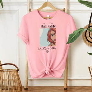 May include: Pink t-shirt with a graphic of a cartoon character and the text "But Daddy I Love Him". The graphic includes a portrait of a character with red hair. The shirt is hanging on a wooden hanger.