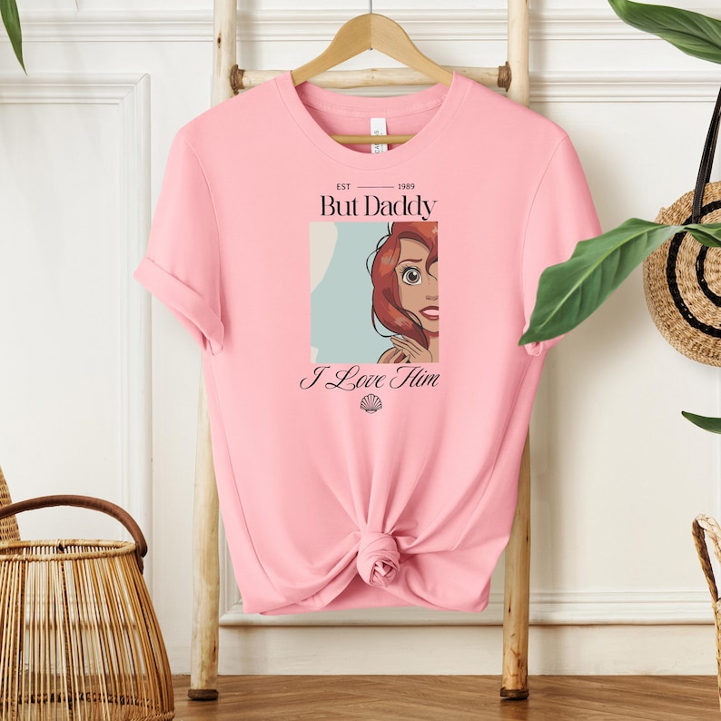May include: Pink t-shirt with a graphic of a cartoon character and the text "But Daddy I Love Him". The graphic includes a portrait of a character with red hair. The shirt is hanging on a wooden hanger.
