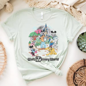 Puede incluir: Camiseta verde claro con un gr&aacute;fico colorido de personajes de Disney, incluyendo Mickey Mouse, Minnie Mouse, Donald Duck y Goofy, frente a un castillo. El texto "Walt Disney World" est&aacute; impreso debajo del gr&aacute;fico.