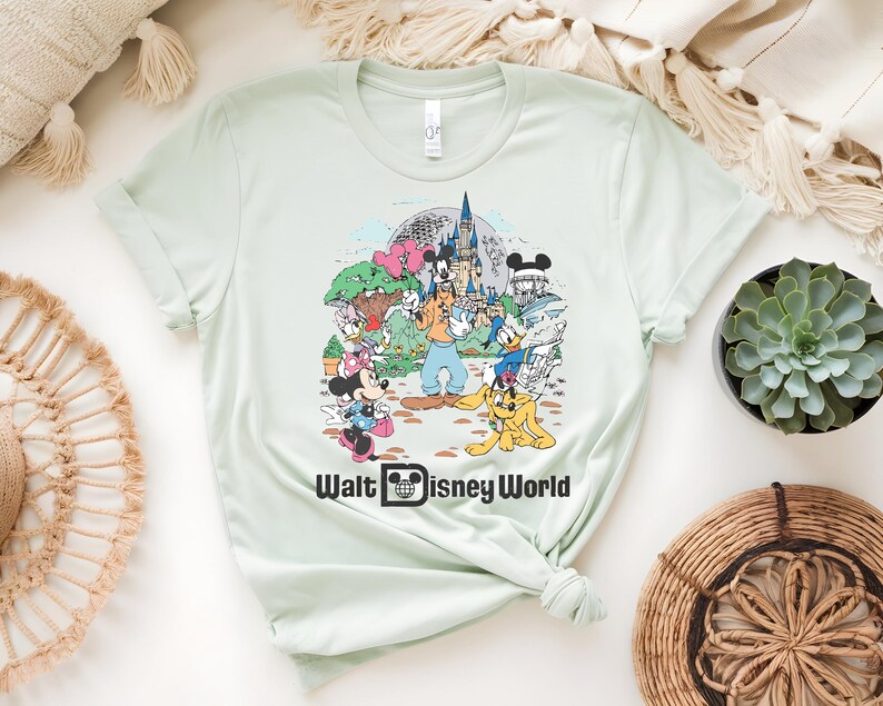 Puede incluir: Camiseta verde claro con un gr&aacute;fico colorido de personajes de Disney, incluyendo Mickey Mouse, Minnie Mouse, Donald Duck y Goofy, frente a un castillo. El texto "Walt Disney World" est&aacute; impreso debajo del gr&aacute;fico.