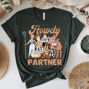 May include: Dark green t-shirt featuring a cartoon graphic of characters with the text "Howdy Partner" in orange. The graphic includes a cartoon of a sheep, a horse, and other characters.