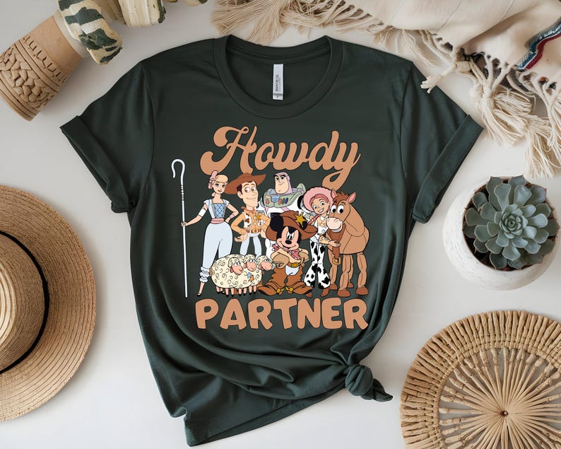 May include: Dark green t-shirt featuring a cartoon graphic of characters with the text "Howdy Partner" in orange. The graphic includes a cartoon of a sheep, a horse, and other characters.