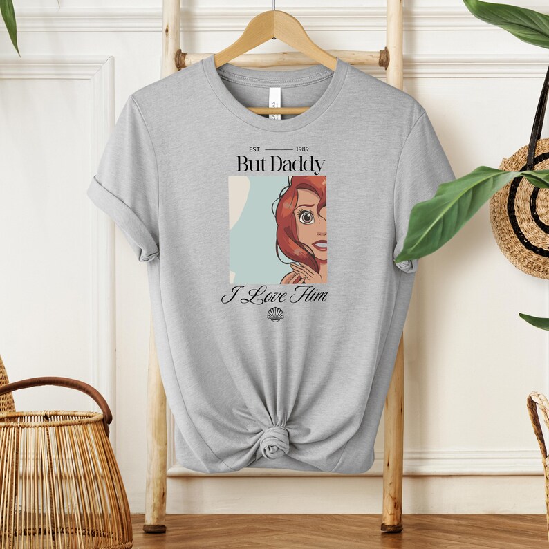 May include: A light gray t-shirt featuring a graphic of a cartoon character with red hair and the text "But Daddy" and "I Love Him". The shirt has a crew neck and short sleeves.