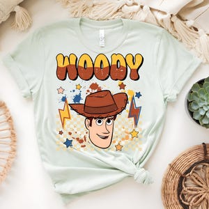 Puede incluir: Camiseta verde claro con un dise&ntilde;o de Woody de Toy Story. La palabra "WOODY" est&aacute; en letras grandes de estilo retro sobre una ilustraci&oacute;n de la cara y el sombrero de Woody. Estrellas y rayos rodean al personaje.