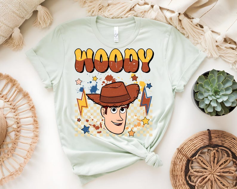 Puede incluir: Camiseta verde claro con un dise&ntilde;o de Woody de Toy Story. La palabra "WOODY" est&aacute; en letras grandes de estilo retro sobre una ilustraci&oacute;n de la cara y el sombrero de Woody. Estrellas y rayos rodean al personaje.