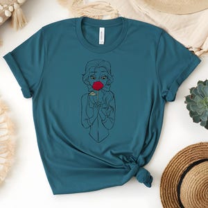 May include: Teal-colored t-shirt featuring a line drawing of a woman wearing a red mask. The graphic is centered on the shirt. The shirt is made of a soft material and has a classic crew neck.
