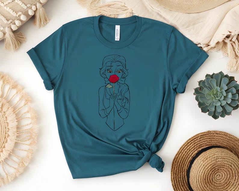 May include: Teal-colored t-shirt featuring a line drawing of a woman wearing a red mask. The graphic is centered on the shirt. The shirt is made of a soft material and has a classic crew neck.