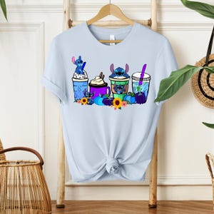 Puede incluir: Camiseta azul claro con un dise&ntilde;o de personajes Stitch en varias bebidas. El dise&ntilde;o incluye elementos azules, morados y amarillos, con girasoles y calabazas. La camiseta est&aacute; colgada en un perchero de madera.