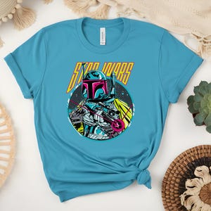 May include: A turquoise t-shirt with a graphic of a Star Wars character in a helmet and holding a weapon, set against a circular space scene. The words "STAR WARS" are above the character in yellow and pink.