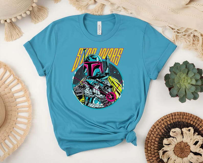 May include: A turquoise t-shirt with a graphic of a Star Wars character in a helmet and holding a weapon, set against a circular space scene. The words "STAR WARS" are above the character in yellow and pink.