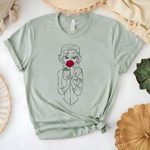 May include: A light green t-shirt featuring a black and white line drawing of a woman holding a red rose to her face. The shirt has a crew neck and short sleeves. The shirt is made of a soft material.