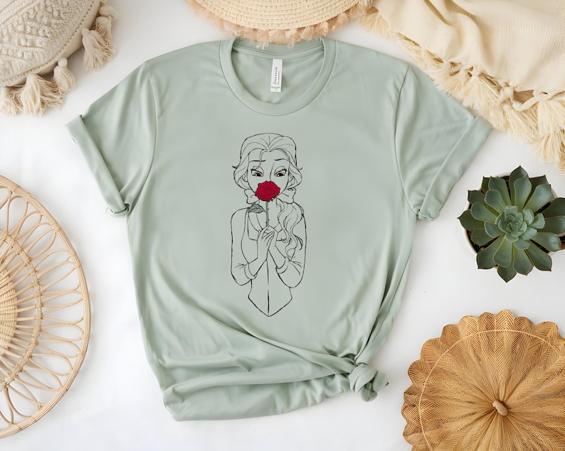 May include: A light green t-shirt featuring a black and white line drawing of a woman holding a red rose to her face. The shirt has a crew neck and short sleeves. The shirt is made of a soft material.