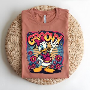 May include: A terracotta-colored t-shirt featuring a cartoon of Daisy Duck with the word "GROOVY" in a colorful, retro font. Daisy is wearing a pink bow and dress, surrounded by flowers and a starburst design.