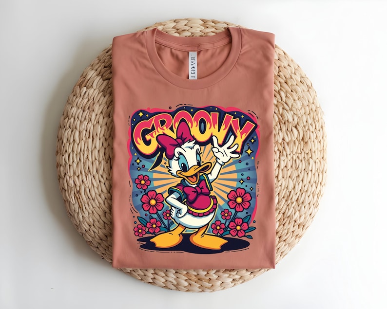 May include: A terracotta-colored t-shirt featuring a cartoon of Daisy Duck with the word "GROOVY" in a colorful, retro font. Daisy is wearing a pink bow and dress, surrounded by flowers and a starburst design.